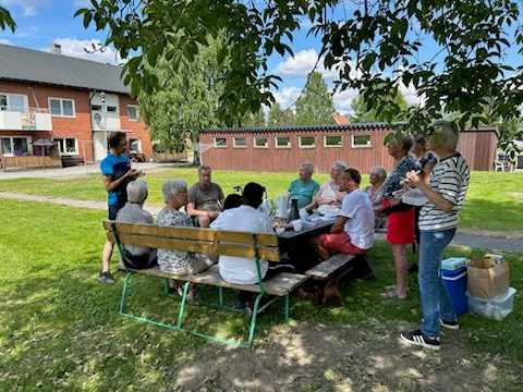 Ett antal personer som sitter runt ett bord under ett trä med ett hyreshus i tegel i bakgrunden